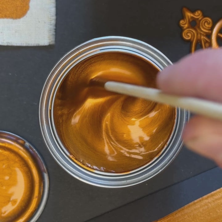 A close-up of Burnt Copper metallic paint being stirred, revealing a deep, fiery orange-gold hue. The paint is swatched onto a dark surface, showing a rich, saturated metallic texture.