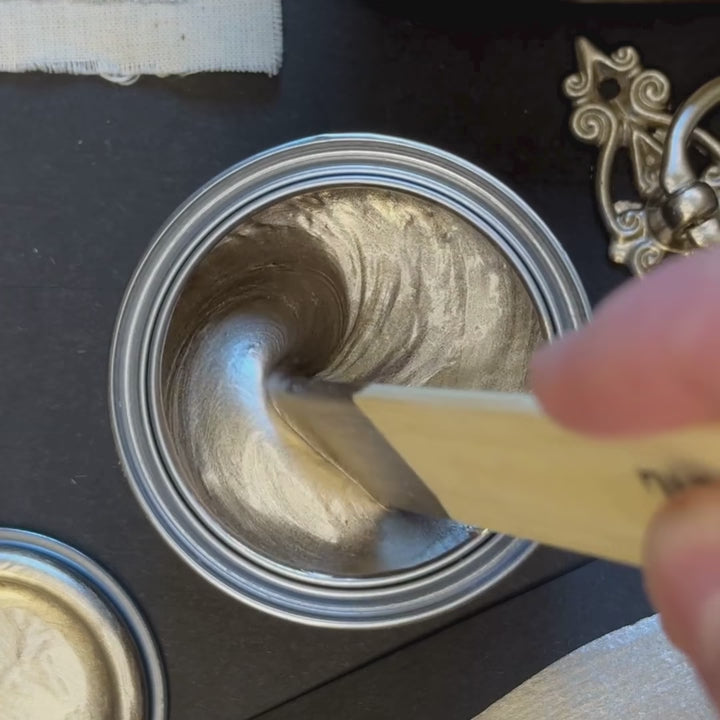 A wooden spatula stirs a thick, cool-toned platinum metallic paint. The paint is then swatched onto a black background with a brush, showing exceptional opacity and a crisp, white-gold metallic brilliance.